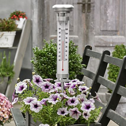 Vonkajší teplomer meria teplotu v stupňoch Celzia a Fahrenheita. Je umiestnený v kvetináči s fialovými petúniami, na záhradnej lavici pred dverami.