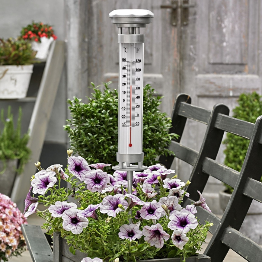 Vonkajší teplomer meria teplotu v stupňoch Celzia a Fahrenheita. Je umiestnený v kvetináči s fialovými petúniami, na záhradnej lavici pred dverami.