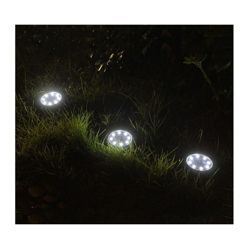 Štyri solárne svietidlá svietia v tráve. Svietidlá sú zapustené do zeme a vyžarujú svetlo. Nachádzajú sa v exteriéri, v nočnom prostredí.