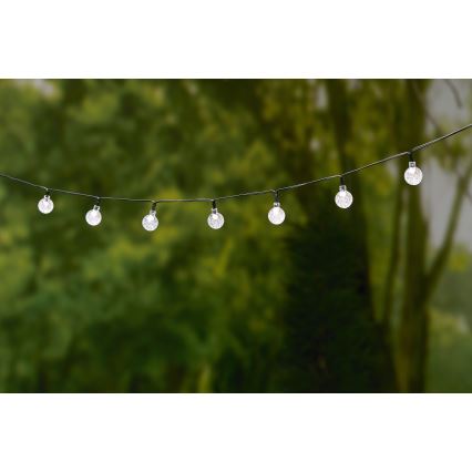 Solárne svetielka visia na šnúre pred rozmazaným pozadím zelenej vegetácie. Guľové sklenené tienidlá sú pripevnené na čiernej šnúre.