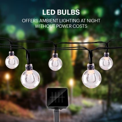 Solárne LED svetielka s guľovitými sklenenými tienidlami svietia v noci. Sú zavesené na čiernom kábli. Solárny panel je umiestnený na zemi. Text: LED BULBS, OFFERS AMBIENT LIGHTING AT NIGHT WITHOUT POWER COSTS.