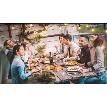 Skupina ľudí posedáva pri dlhom stole na terase a spoločne večeria. Jedia, pijú víno a rozprávajú sa. Nad stolom sú zavesené svetielka.