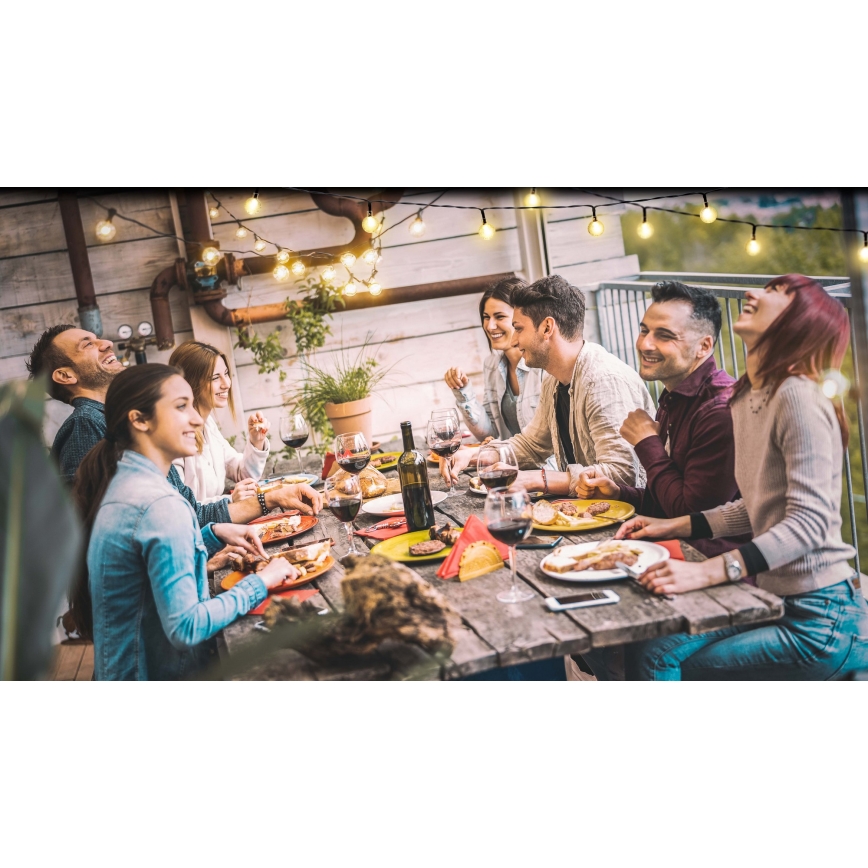 Skupina ľudí posedáva pri dlhom stole na terase a spoločne večeria. Jedia, pijú víno a rozprávajú sa. Nad stolom sú zavesené svetielka.
