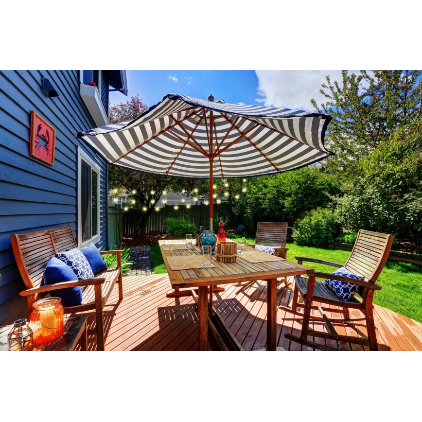 Vonkajší jedálenský stôl so stoličkami a lavicami pod pruhovaným slnečníkom na drevenom nábytku. Na stole sú taniere a dekorácie. Okolitá záhrada s bujnou zeleňou.