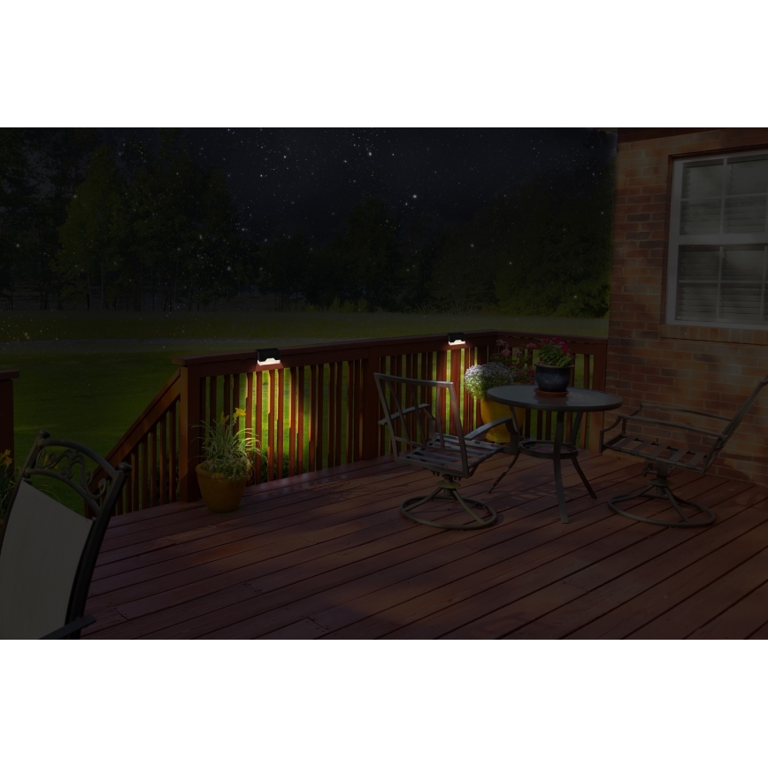 Solárne svietidlá osvetľujú drevenú terasu v noci. Svietidlá sú namontované na zábradlí terasy. Na terase sa nachádza vonkajší jedálenský set s dvoma stoličkami a stolom.