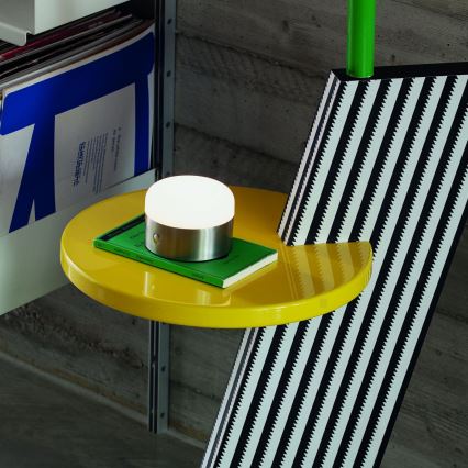 Malá stolová lampa svieti na malej žltej odkladacie ploche. Plocha je súčasťou väčšej, čierno-bielo pruhovanej konštrukcie. Vedľa lampy leží zelená kniha.