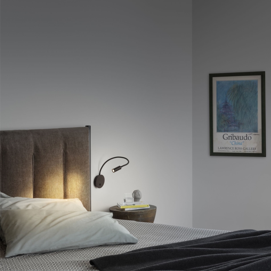 Nástenná lampa s flexibilným ramenom svieti v spálni. Lampa je pripevnená k čelu postele. Na nočnom stolíku sú kniha a pohárik. Na stene visí obraz s textom: Gribaudo „China“ LAWRENCE ROSS GALLERY.