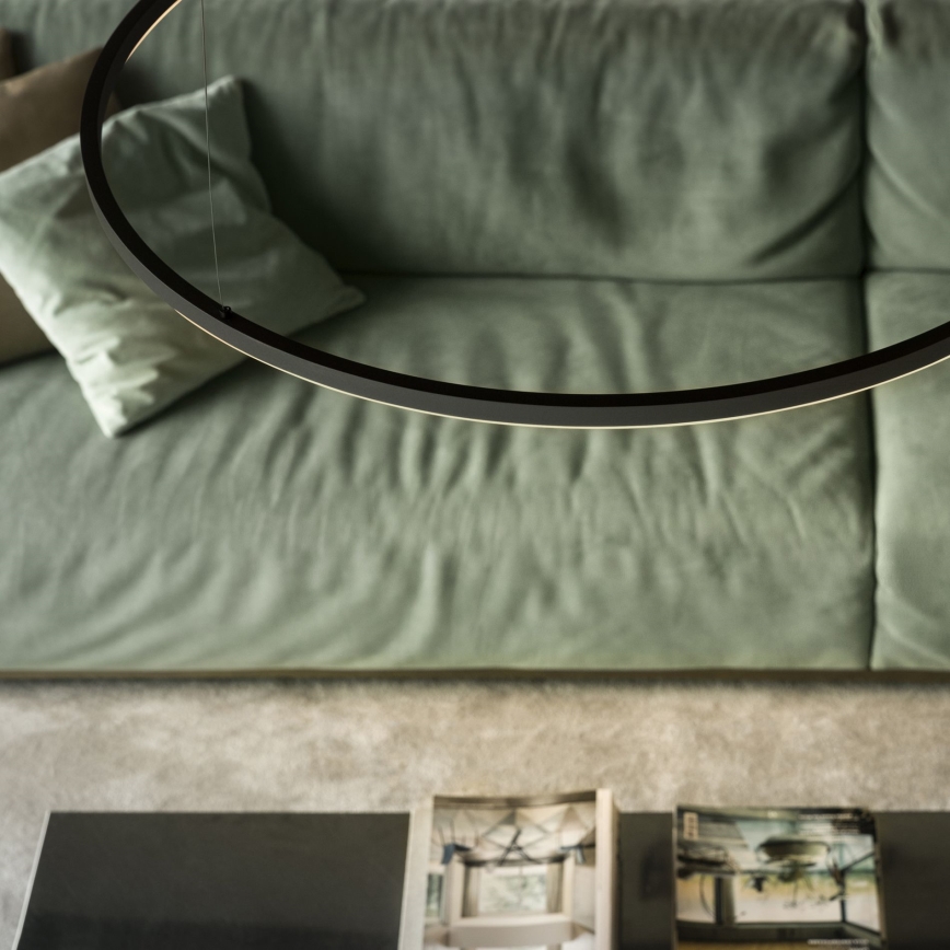 Kruhové svietidlo visí nad svetlozelenou sedačkou v obývacej izbe. Pod svietidlom sa nachádza konferenčný stolík s časopismi.