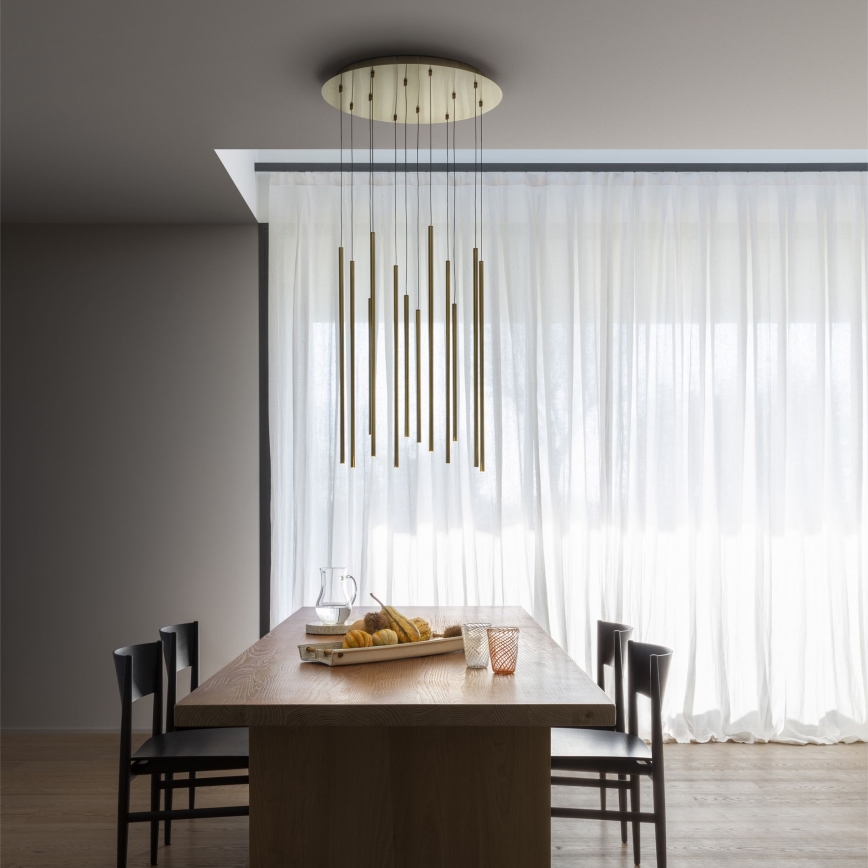Lustr s viacerými zlato-medenými tyčinkami visí nad dreveným jedálenským stolom. Na stole je ovocie, nádoba a poháre. Stojí tam štyri stoličky a v pozadí sú svetlé závesy.