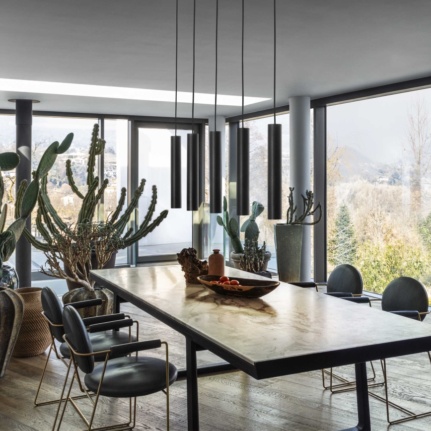 Dlhý jedálenský stôl z svetlého kameňa stojí v priestrannej miestnosti s veľkými oknami. Nad stolom visí päť dlhých čiernych závesných svietidiel. V pozadí sú viditeľné rastliny v kvetináčoch.