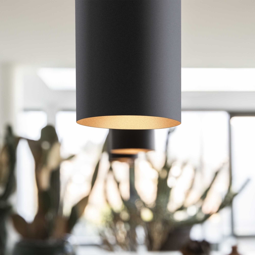 Cylindrická závesná lampa s tmavosivým povrchom a zlatým vnútorným okrajom visí v priestore s rozmazaným pozadím rastlín.