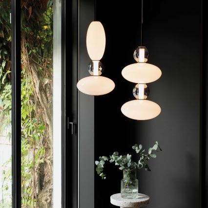 Dve závesné lampy s oválnymi tienidlami a kovovými prvkami visia v tmavej miestnosti pri okne s výhľadom na zeleň. Na malom stolíku pod lampami je váza s vetvičkami.