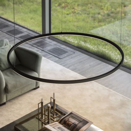 Kruhové svietidlo visí v interiéri nad sedačkou a kávovým stolíkom s knihou a sviečkami. Svetlo je umiestnené v blízkosti veľkého okna s výhľadom do zelene.