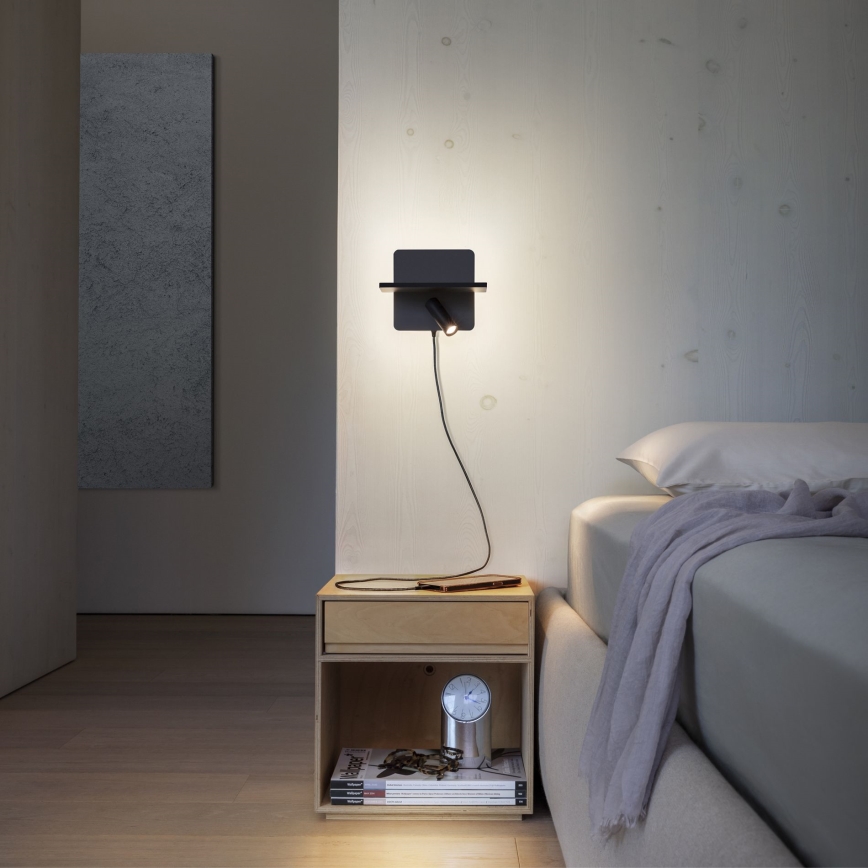 Nástená lampa so vstavanou poličkou svieti v spálni. Kábel lampy je zapojený do zásuvky na nočnom stolíku, na ktorom sa nachádzajú časomiera a časopisy.