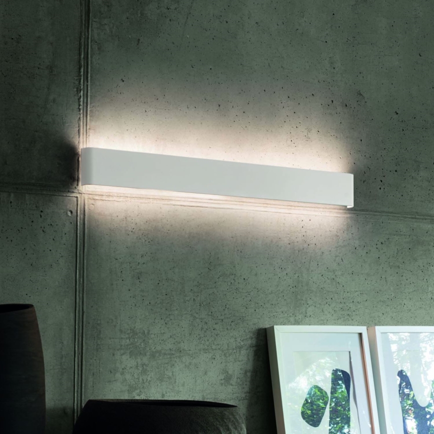 Dlhá biela nástenná lampa svieti na betónovej stene. Lampa je umiestnená nad dvoma obrázkami v rámčekoch.