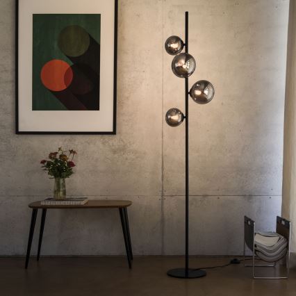 Stojacia lampa s čiernym stojanom a štyrmi sklenenými guľami svieti v miestnosti s betónovou stenou. Popri nej sa nachádza nízky konferenčný stolík a obraz s abstraktnými kruhmi.