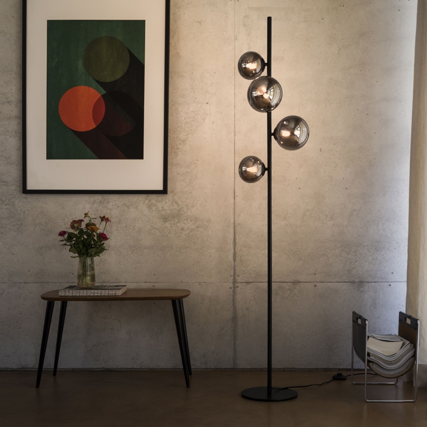 Stojacia lampa s čiernym stojanom a štyrmi sklenenými guľami svieti v miestnosti s betónovou stenou. Popri nej sa nachádza nízky konferenčný stolík a obraz s abstraktnými kruhmi.