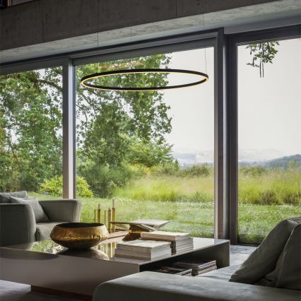 Okrúhle závesné svietidlo svieti v interiéri. Nachádza sa nad nízkou sedačkou a konferenčným stolíkom, na ktorom sú knihy a miska. Za nimi je veľké okno s výhľadom na lúku a stromy.