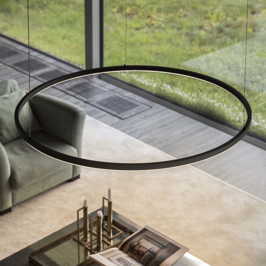 Kruhové svietidlo visí nad sedačkou a konferenčným stolíkom v interiéri. Svietidlo je čierne, osvetľuje miestnosť. V pozadí je vidieť veľké okno s výhľadom na zeleň.