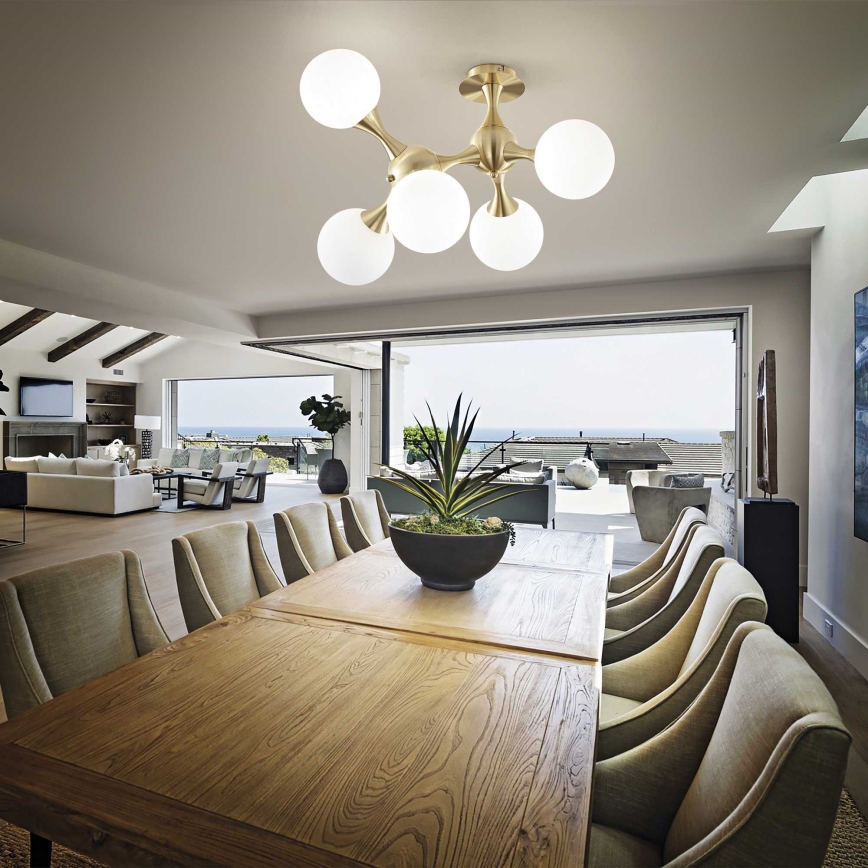 Lustro s piatimi bielymi guľovými tienidlami visí nad dreveným jedálenským stolom s mnohými stoličkami. Stôl stojí v jedálni s výhľadom na oceán cez otvorené dvere.