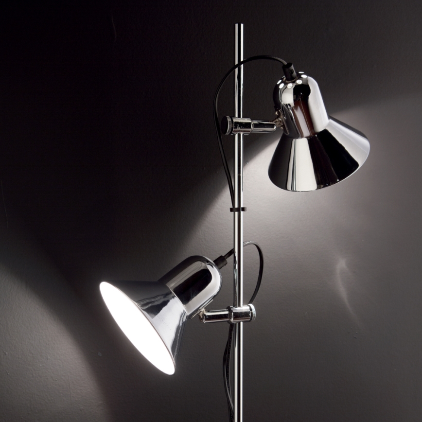 Stojacia lampa s dvoma chrómovými tienidlami svieti v tmavej miestnosti. Tienidlá sú na nastaviteľných ramenách.