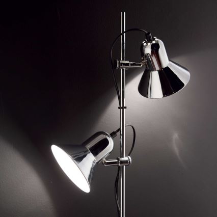Stojacia lampa s dvoma chrómovanými ramenami a tienidlami smeruje svetlo do priestoru. Lampa je umiestnená pri tmavej stene.