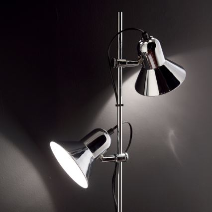Stojacia lampa s dvoma chrómovými tienidlami svieti v tmavej miestnosti. Tienidlá sú na nastaviteľných ramenách.