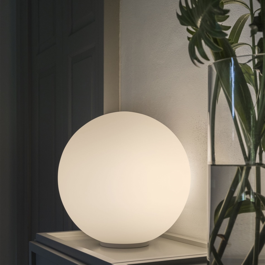 Guľová stolová lampa svieti. Nachádza sa na bielom stolíku pri stene, vedľa vázy s rastlinou.
