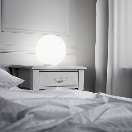Guľatá stolová lampa svieti na nočnom stolíku. Stolík stojí v spálni pri posteli s bielou posteľnou bielizňou a blízko bielych závesov.