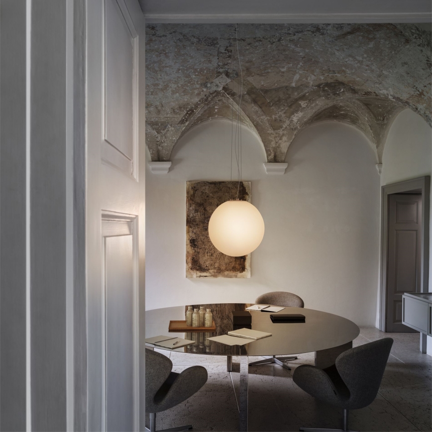 Guľová lampa svieti nad okrúhlym stolom s dokumentmi a stoličkami v miestnosti s klenutým stropom a obrazom na stene.
