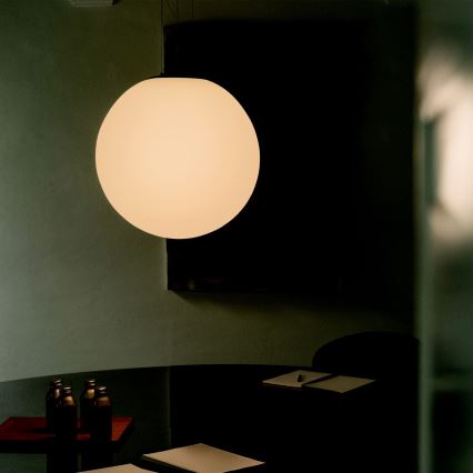 Guľová lampa svieti v tmavej miestnosti. Na stole pod lampou sú tri fľaše a zápisníky. Stena je tmavozelená.