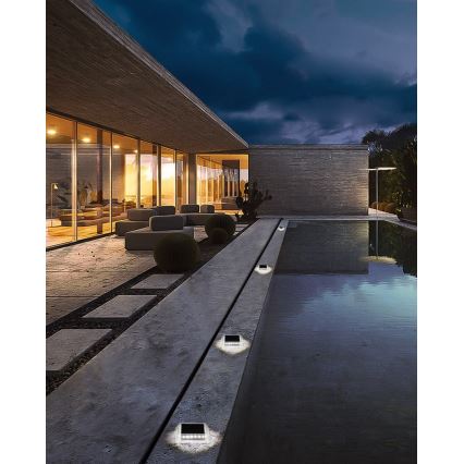 Solárne svietidlá osvetľujú chodník vedľa bazéna pri modernom dome. Svietidlá sú zabudované do dlažby. V pozadí je vidieť modernú architektúru a záhradu.