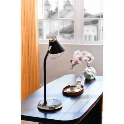 Stolová lampa s čiernym tienidlom a zlatým detailom stojí na tmavom stole. Vedľa nej je váza s bielymi orchideami a tácka s šálkou. Pozadie tvorí okno s výhľadom na mesto.