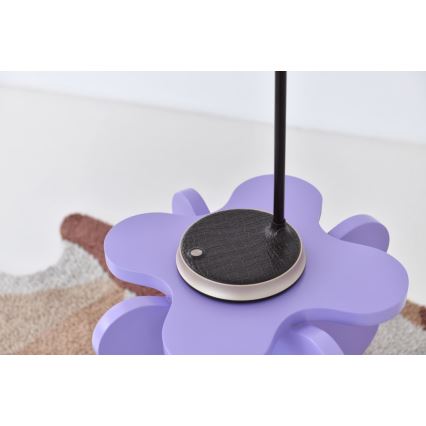 Stolová lampa s čiernou základňou a kovovou tyčou je umiestnená na fialovej podložke v tvare kvetu. Podložka stojí na koberci s hnedými a sivými odtieňmi.