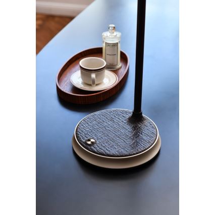 Stolová lampa stojí na tmavom stole. Jej základňa je pokrytá hnedou kožou s imitáciou krokodílej kože. V blízkosti sa nachádza oválny drevený podnos s hrnčekom a malou fľaškou.