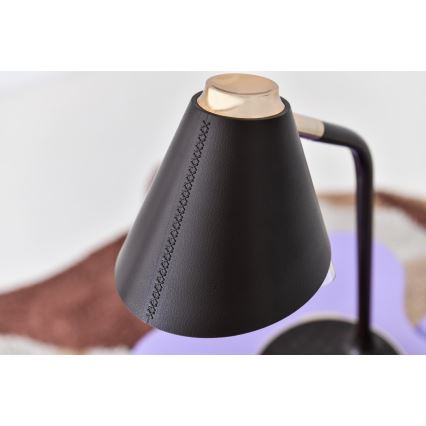 Stolová lampa s kužeľovitým tienidlom z tmavej kože, ozdobeným stehom, a zlatou kovovou časťou na vrchu, stojí na fialovom podklade. Lampa je umiestnená na hnedom koberci.