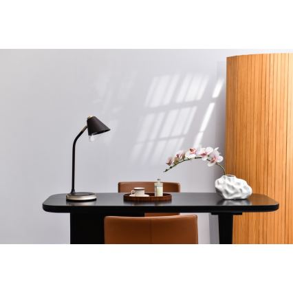 Čierna stolová lampa s kužeľovitým tienidlom stojí na čiernom stole. Na stole je tiež podnos s hrnčekom a sviečkou, a váza s umelou orchideou. Vedľa stola je drevená priečka.
