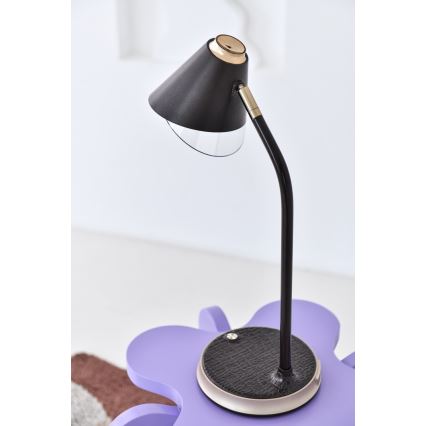Stolová lampa s čiernym tienidlom a kovovou nohou stojí na fialovom stole. Lampa má na spodku okrúhlu, tmavosivú podložku. Na stole je tiež časť hnedého koberca.