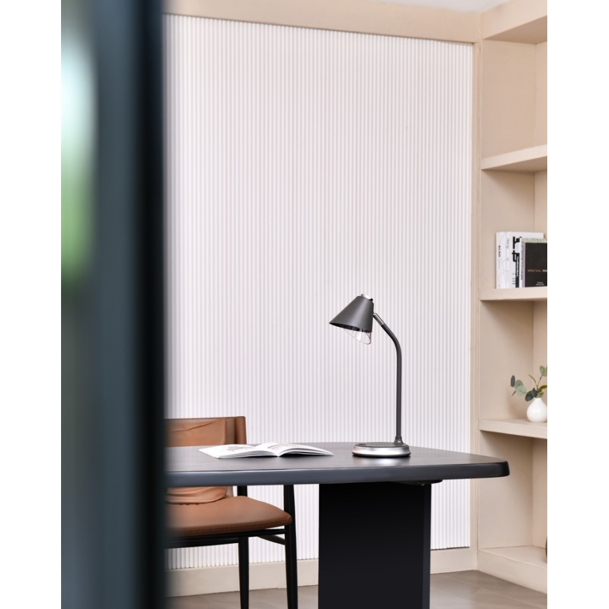Stolová lampa stojí na tmavom stole v pracovni. Lampa s kuželovitým tienidlom osvetľuje otvorenú knihu. V pozadí je vidieť polica s knihami a stena s vertikálnymi líniami.