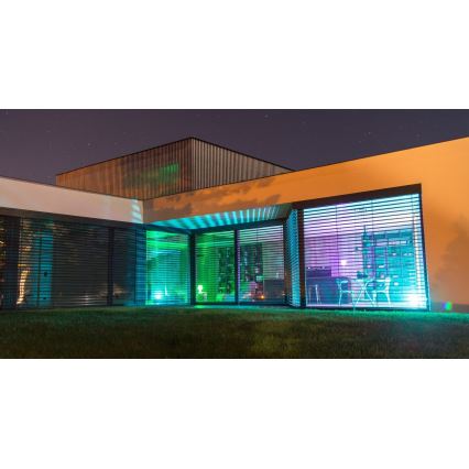 Moderná budova s veľkými oknami a žalúziami v noci. Vnútorné osvetlenie je farebné, modro-zelené. Vonkajšie osvetlenie je tiež farebné, tráva je zelená.