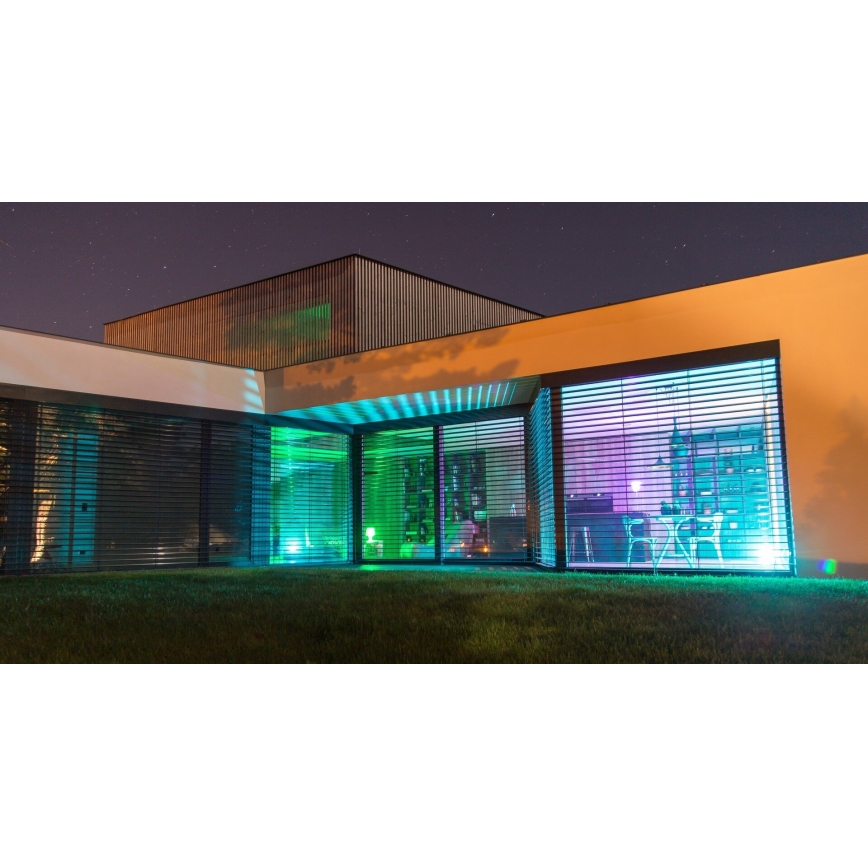 Moderná budova s veľkými oknami a žalúziami v noci. Vnútorné osvetlenie je farebné, modro-zelené. Vonkajšie osvetlenie je tiež farebné, tráva je zelená.