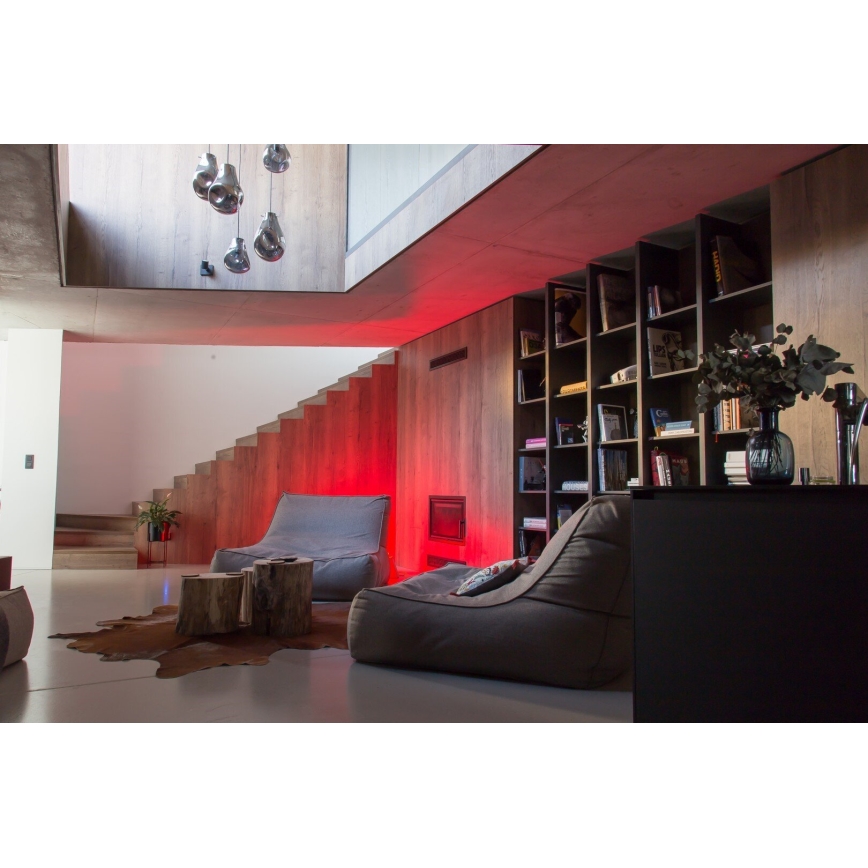 Veľká hnedá sedačka s vankúšom je umiestnená v priestrannej miestnosti s drevenou knižnicou, schodiskom a červeným osvetlením. Na podlahe je kožušina a drevený stolík.