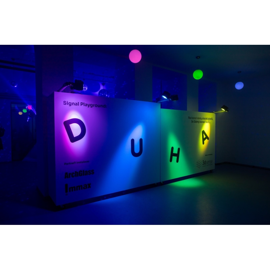 Veľké písmená D, U, H, A sú podsvietené farebnými svetlami na bielych paneloch. Na paneloch je text: Signal Playground, ArchGlass, Immax, The iconic duality staple evolved to building homes next. Miestnosť je slabo osvetlená, na strope visia farebné gule.
