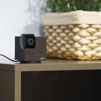 Webkamera značky EMOS stojí na sivom nábytku. Je pripojená káblom. Za ňou je košík s izbovou rastlinou.