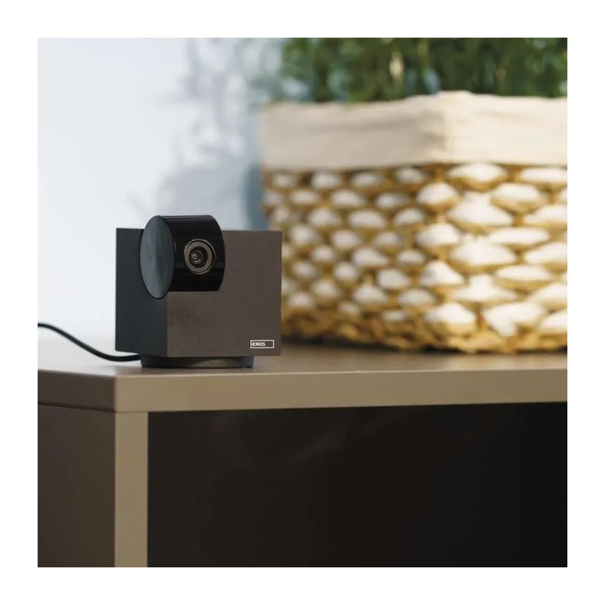 Webkamera značky EMOS stojí na sivom nábytku. Je pripojená káblom. Za ňou je košík s izbovou rastlinou.