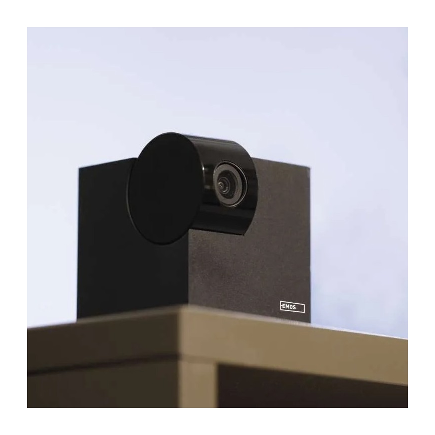 Čierna kamera EMOS je umiestnená na tmavosivej poličke. Kamera je otočená dopredu a sníma obraz. Polica je súčasťou nábytku.