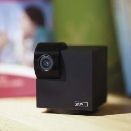 Čierna webkamera tvaru kocky s nápisom „EMOS“ stojí na drevenom povrchu. Objektív kamery je smerovaný dopredu. Na pozadí je rozmazané prostredie s knihou a časťou osoby.