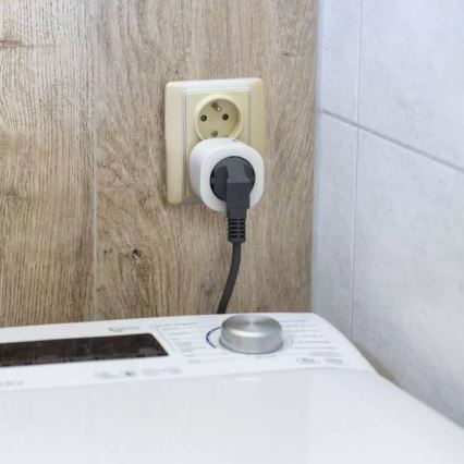 Smart zástrčka je zapojená do elektrickej zásuvky na stene. Kábel vedie k práčke stojacej na podlahe v kúpeľni.