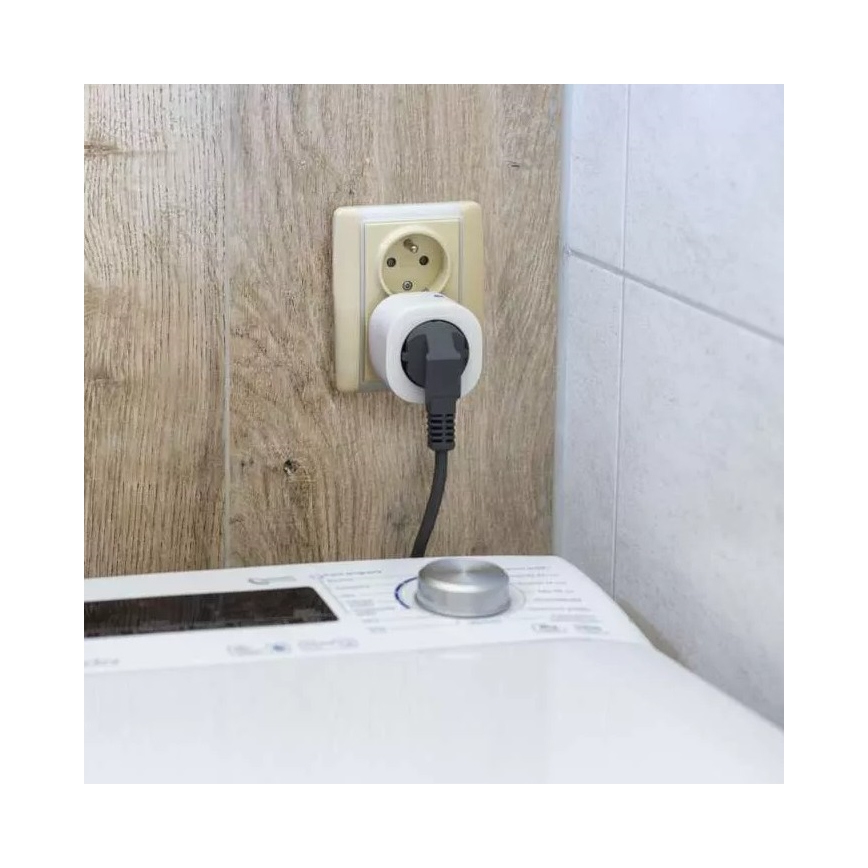 Smart zástrčka je zapojená do elektrickej zásuvky na stene. Kábel vedie k práčke stojacej na podlahe v kúpeľni.