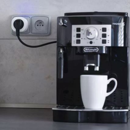 Espresso automat Delonghi Magnifica S je zapojený do inteligentnej zásuvky. Kávovar pripravuje kávu do bieleho hrnčeka. Celá scéna sa odohráva na kuchynskej linke.